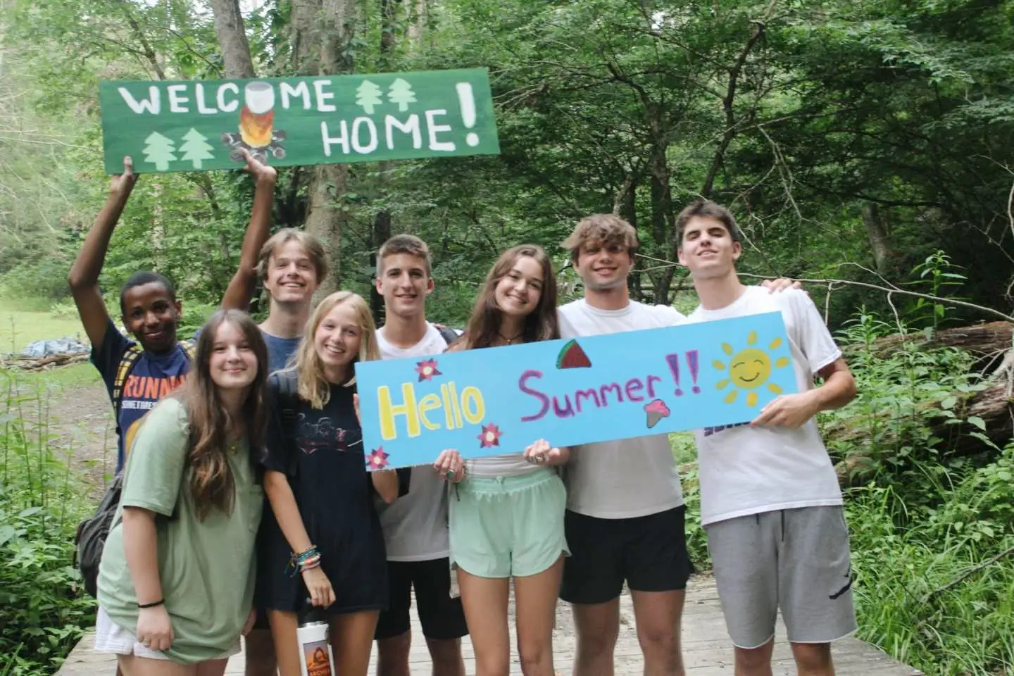 A co-ed group of Counselors in Training hold a "Welcome" sign and a "Hello Summer" sign