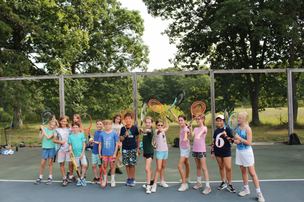 A group of campers stand together on a tennis court. Some hold rackets